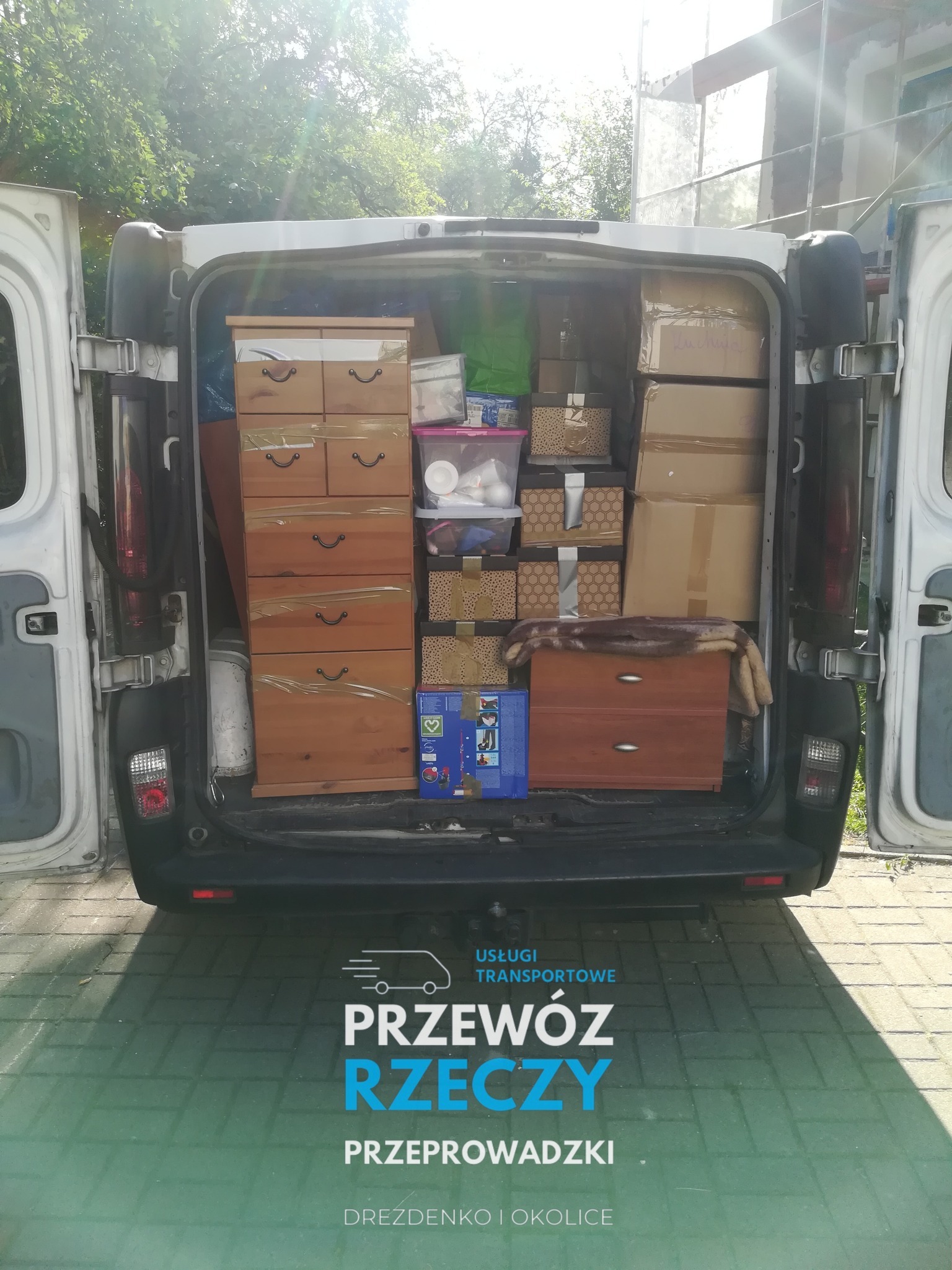 Wnętrze bagażnika białego busa wypełnione meblami, kartonami i pojemnikami podczas słonecznego dnia, przygotowane do transportu, z widocznym logo firmy na dole zdjęcia.