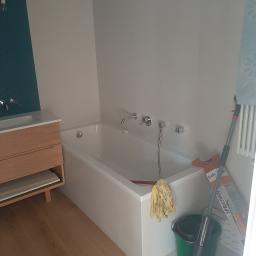 Damian Chojnacki - Nowoczesna łazienka z drewnianą szafką pod umywalką, białą wanną i mopem SprayMop w rogu, prawdopodobnie po sprzątaniu.