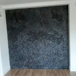 Ściana wykończona dekoracyjną okładziną imitującą kamień w odcieniach szarości i czerni, z widoczną strukturą i fugami, w pomieszczeniu z drewnopodobną podłogą.