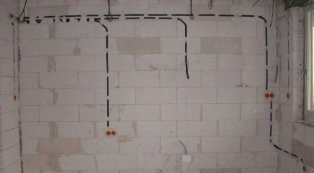 Ściana z betonu komórkowego z wyfrezowanymi kanałami pod instalację elektryczną, widoczne przewody i puszki elektryczne w kolorze pomarańczowym, okno w prawym górnym rogu.