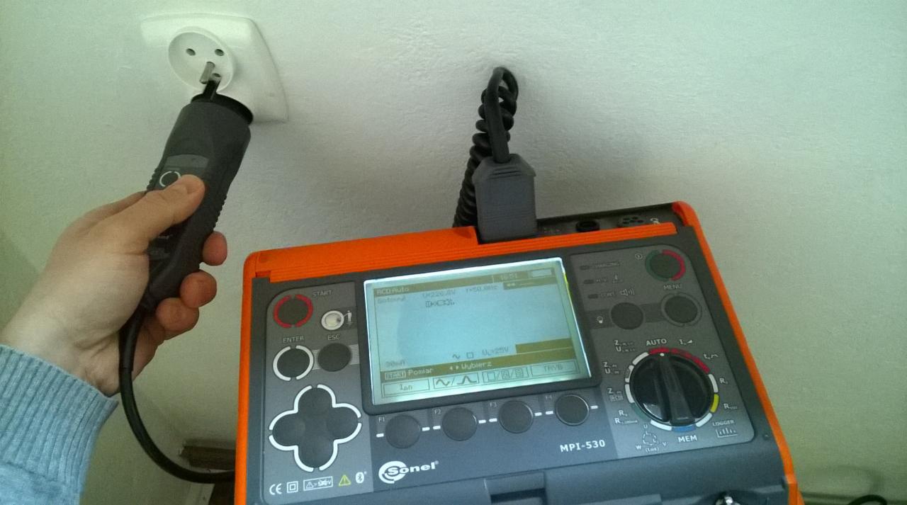 Elektryk mierzy napięcie w gniazdku za pomocą miernika Sonel MPI-530, widoczny przewód pomiarowy wpięty do gniazdka.