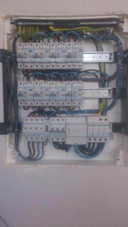 Otwarta skrzynka bezpiecznikowa z widocznymi bezpiecznikami, przewodami w kolorach niebieskim, żółtym i czarnym oraz listwami montażowymi wewnątrz wnęki w ścianie.