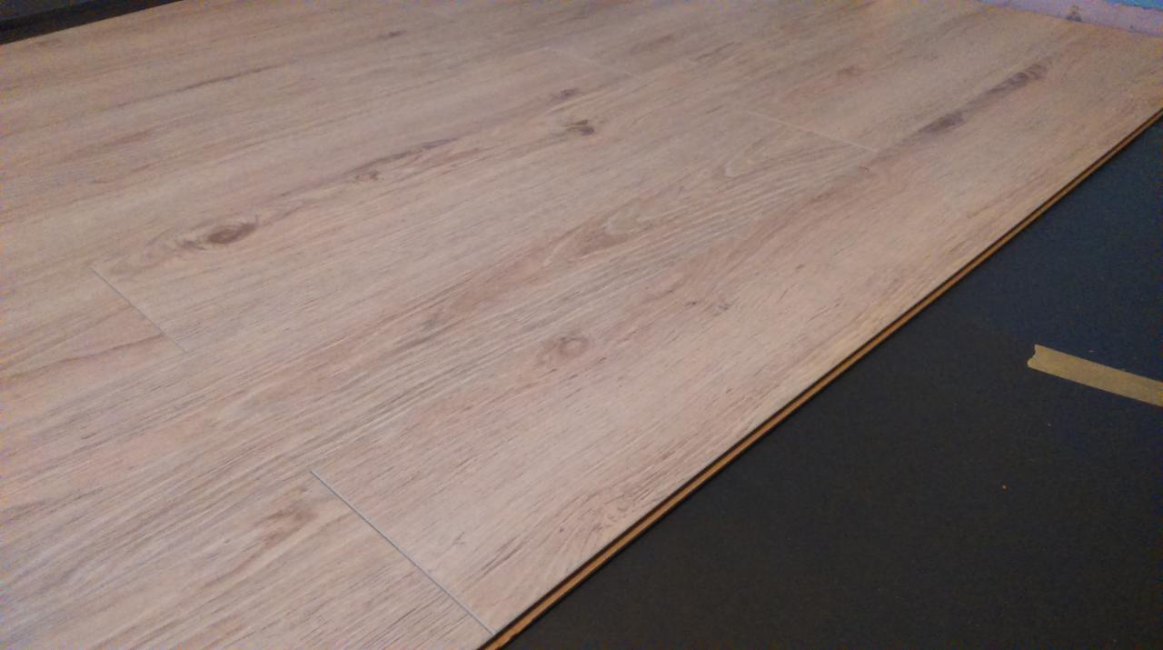 Jasne panele podłogowe ułożone na czarnym podkładzie, widoczny fragment taśmy klejącej.