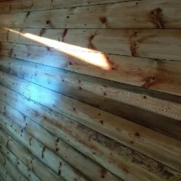 Ściana wyłożona jasnymi, drewnianymi deskami z widocznymi słojami i sękami, oświetlona ukośnym promieniem słońca tworzącym jasną smugę.