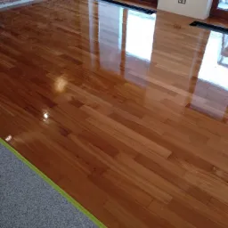 Świeżo polakierowana, błyszcząca podłoga z parkietu w odcieniach brązu, z widoczną taśmą malarską zabezpieczającą krawędzie przy szarej posadzce. W tle ściana z cegły i okno.