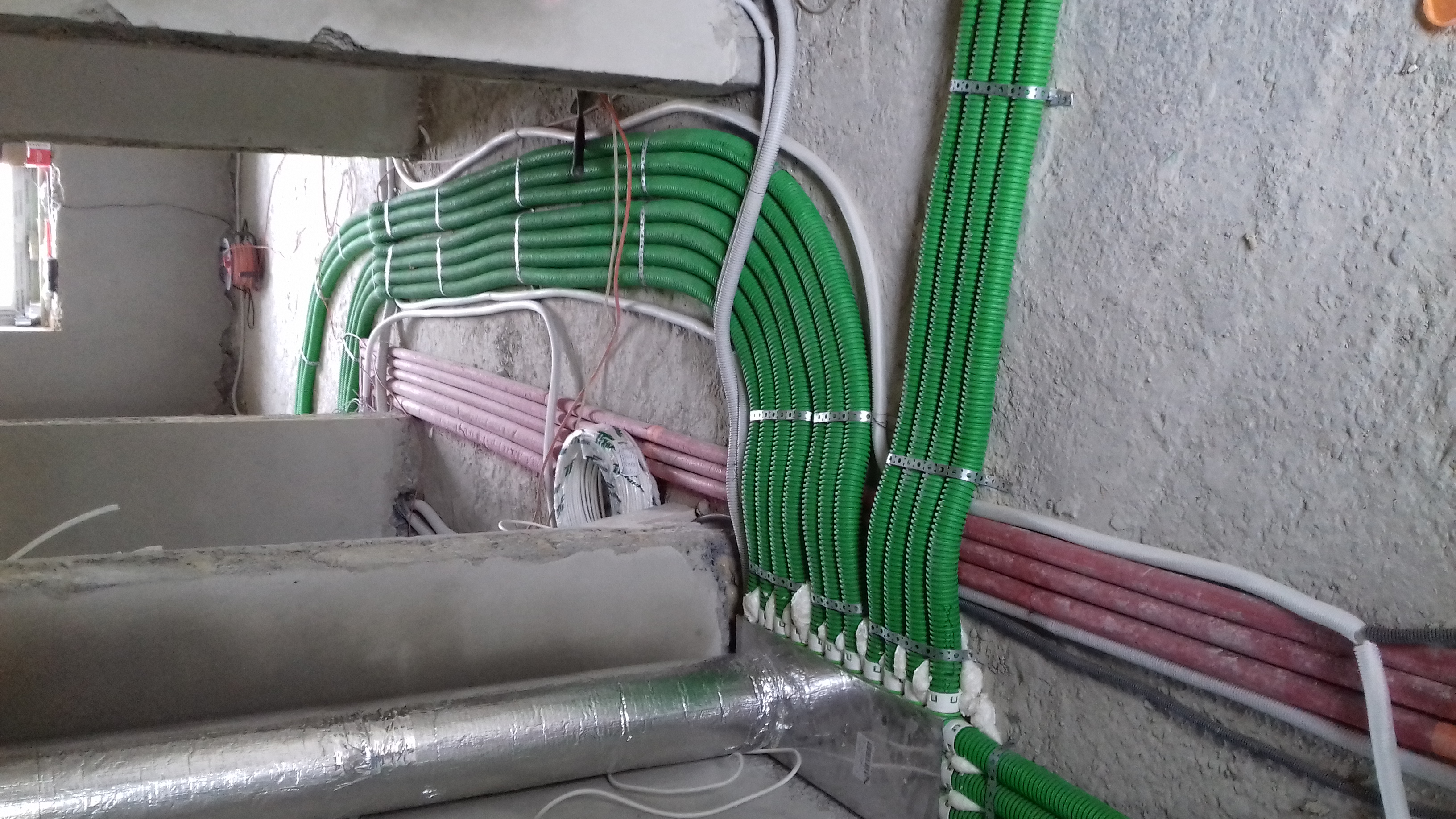 Rzędy zielonych i różowych rur wentylacyjnych, zabezpieczonych metalowymi obejmami, biegnące wzdłuż surowej betonowej ściany w budynku w trakcie budowy.