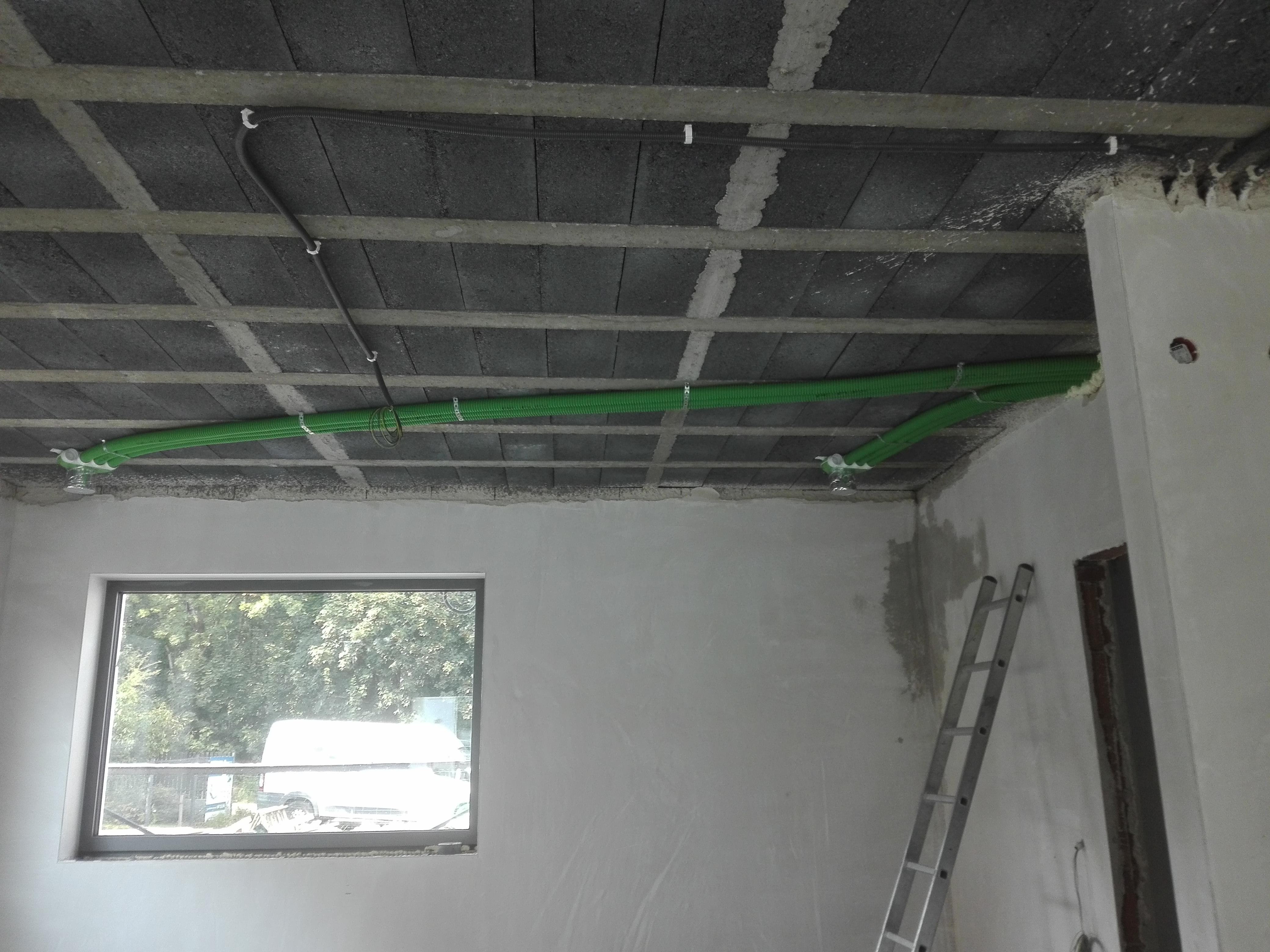 Zielone rury wentylacyjne przymocowane do sufitu z betonowych bloczków w surowym stanie, widoczne okno i drabina.