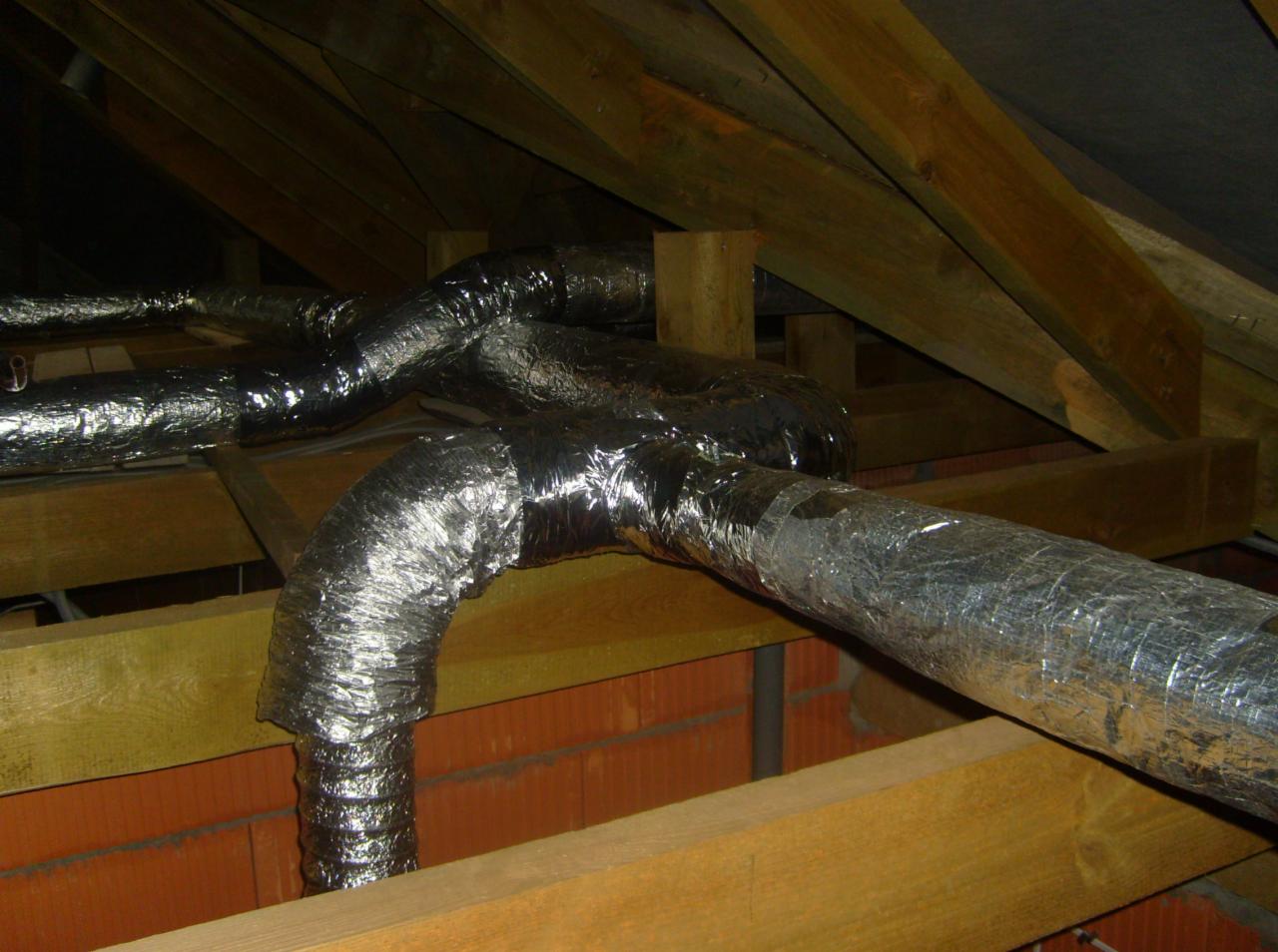 Izolowane, elastyczne przewody wentylacyjne z folii aluminiowej, poprowadzone w przestrzeni poddasza z drewnianą konstrukcją i fragmentem muru z cegły.