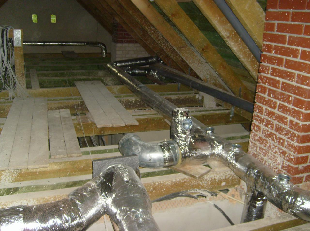 Instalacja przewodów wentylacyjnych w przestrzeni poddasza z widocznymi elementami konstrukcji drewnianej i fragmentem komina z cegły.