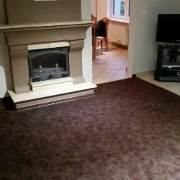 Brązowa wykładzina dywanowa w salonie z kominkiem i telewizorem, widok na płytki w przejściu do innego pomieszczenia.