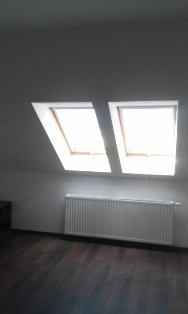 Poddasze z dwoma oknami dachowymi, ciemna drewniana podłoga i biały grzejnik pod oknami.