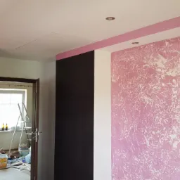 Pokój po odświeżeniu ścian: różowa ściana z efektem dekoracyjnym, widoczna drabina i akcesoria malarskie w tle.