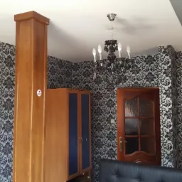 Pokój z tapetą w czarno-białe ornamenty, drewnianą szafą i drzwiami, oraz czarnym żyrandolem.