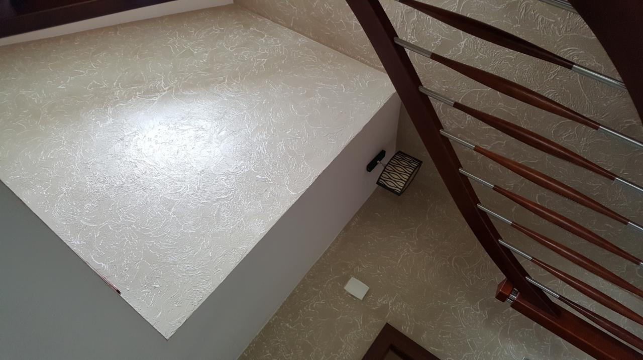 Ściana i sufit w klatce schodowej z dekoracyjną strukturą, widoczna lampa ścienna i fragment drewnianej balustrady.