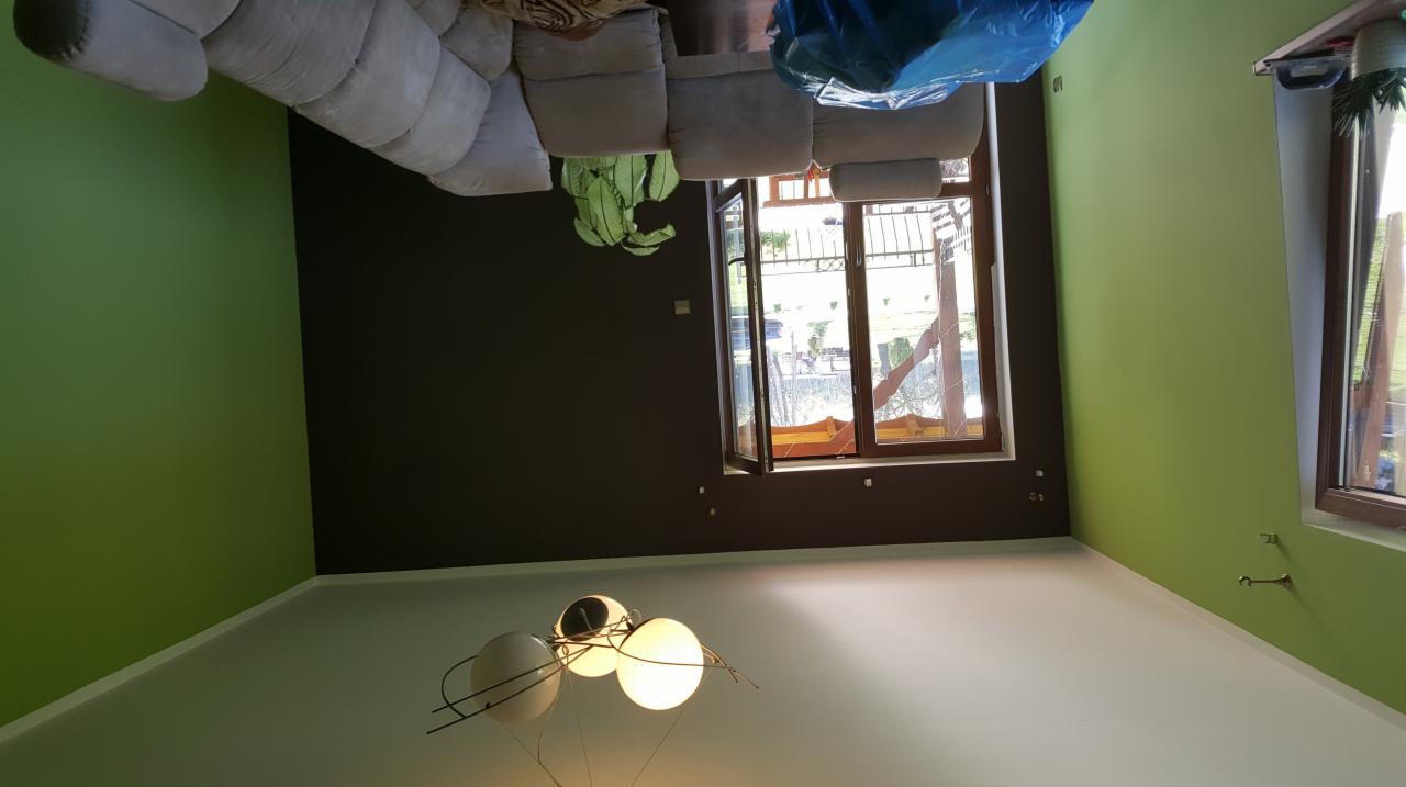 Pomalowane wnętrze pokoju w dwóch kolorach: zielonym i ciemnobrązowym. Widoczny fragment sofy, okno i nowoczesna lampa.