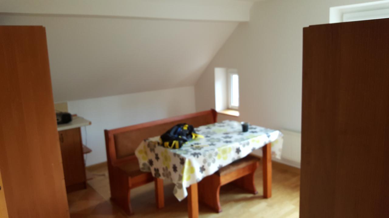 Pokój na poddaszu ze skośnym sufitem, drewnianym stołem z ławami i oknem w ścianie szczytowej, widoczne elementy wyposażenia kuchni w tle.