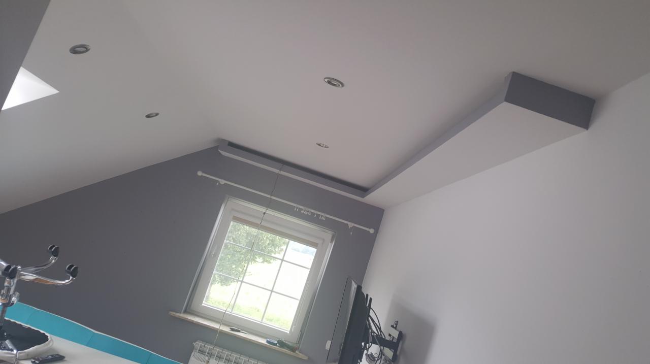 Pokój na poddaszu z szarą ścianą, białym sufitem z oświetleniem punktowym i oknem. Widoczny fragment biurka z krzesłem.