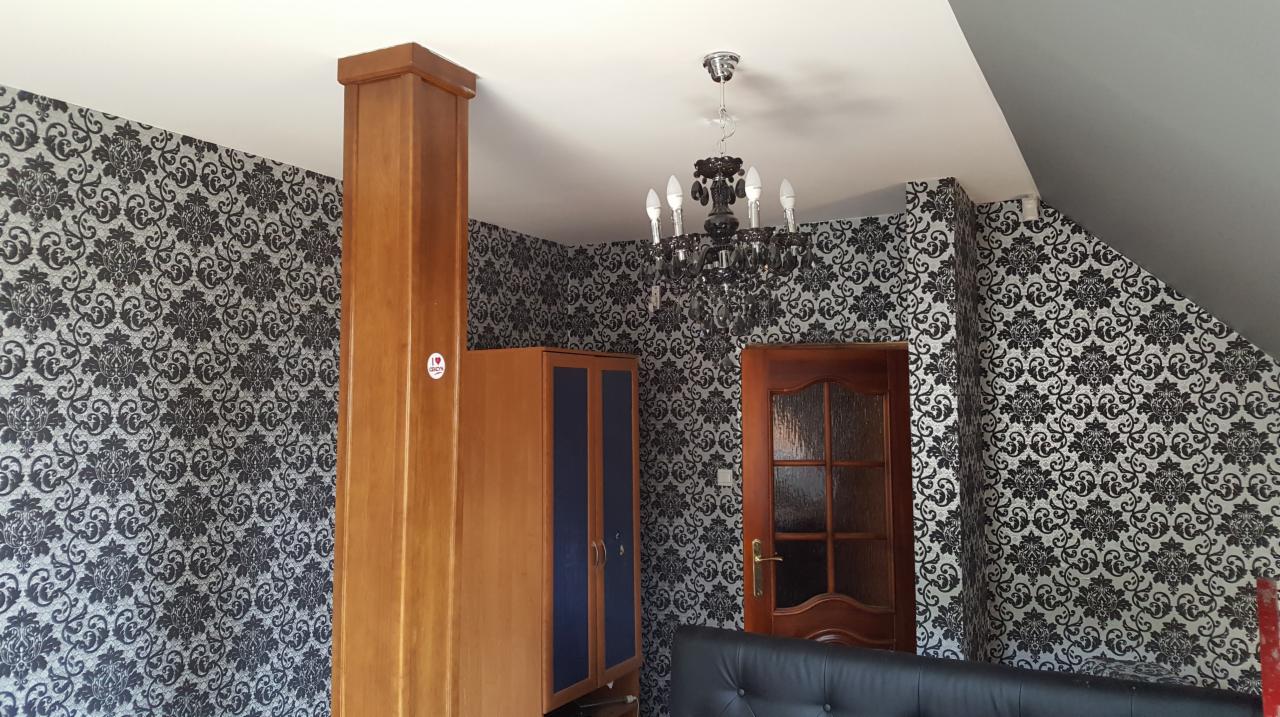 Pokój z tapetą w czarno-białe ornamenty, drewnianą szafą i drzwiami, oraz czarnym żyrandolem.