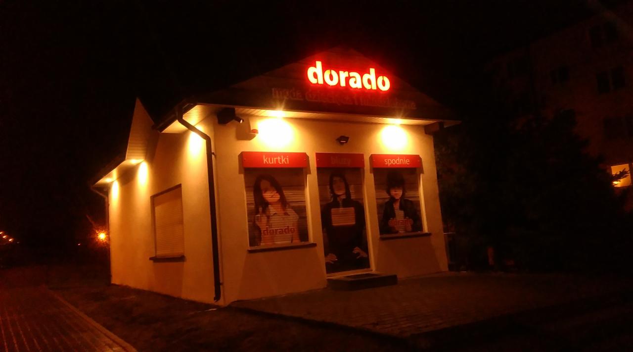 Nocny widok na oświetlony budynek sklepu odzieżowego 'dorado' z trzema oknami wystawowymi prezentującymi plakaty z ofertą: kurtki, bluzy i spodnie.