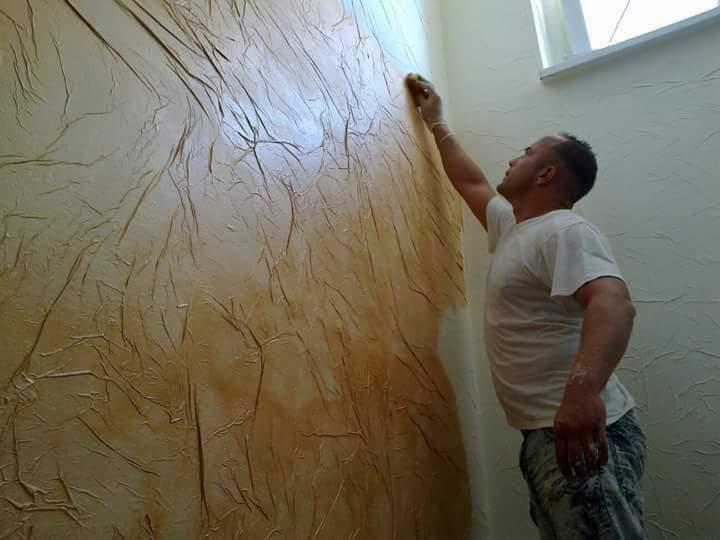 Mężczyzna w rękawicy aplikuje gąbką dekoracyjną farbę strukturalną w odcieniach beżu i brązu na ścianę w pomieszczeniu.
