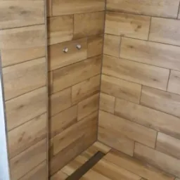 Kabina prysznicowa wykończona płytkami imitującymi drewno, z odpływem liniowym w podłodze i chromowanymi punktami mocowania.