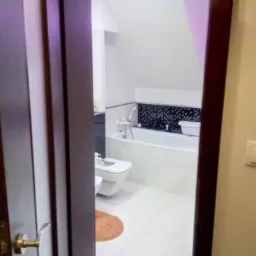 Widok na białą łazienkę z wanną, bidetem i toaletą przez uchylone, ciemne drzwi. Na podłodze widoczny pomarańczowy dywanik.