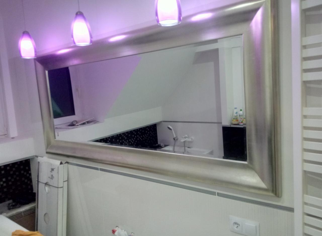 Duże lustro w srebrnej ramie odbija wnętrze wyremontowanej łazienki z czarno-białą mozaiką nad wanną i trzema lampami sufitowymi o fioletowym świetle.