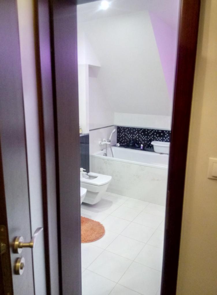 Widok na białą łazienkę z wanną, bidetem i toaletą przez uchylone, ciemne drzwi. Na podłodze widoczny pomarańczowy dywanik.