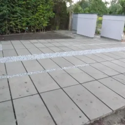 Szare płyty chodnikowe ułożone w regularny wzór, oddzielone pasami z jasnej kostki granitowej, z widocznymi elementami zieleni w tle.