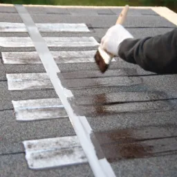 Malowanie papy na dachu, widoczna biała taśma uszczelniająca i pędzel w rękawicy ochronnej, fragment pomalowanej powierzchni.