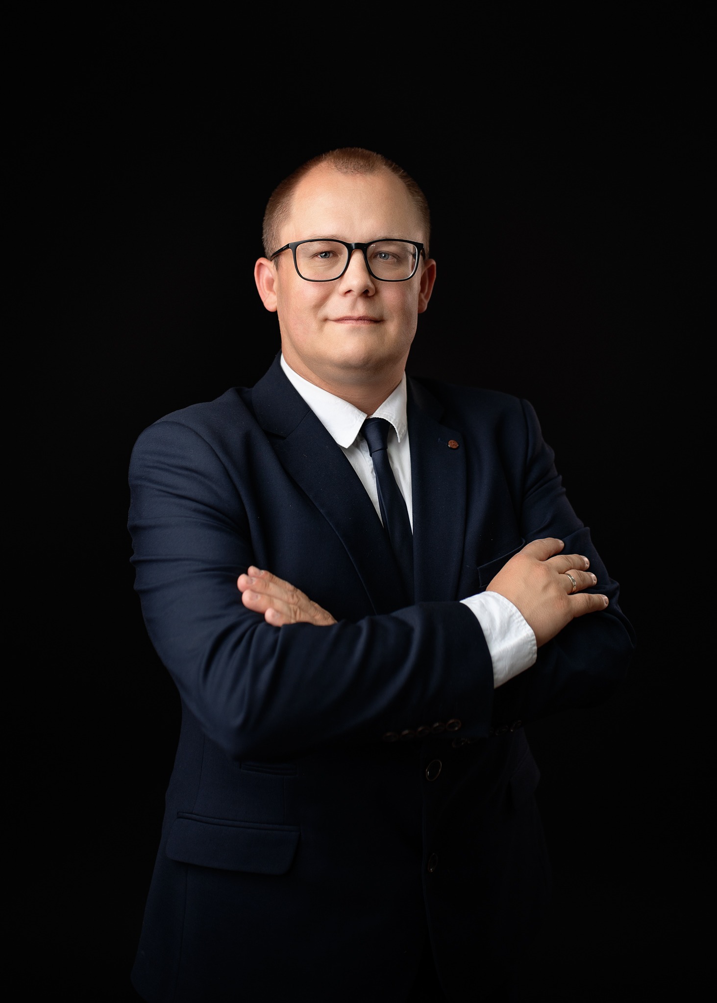 Portret mężczyzny w okularach i granatowym garniturze na czarnym tle, z rękami skrzyżowanymi na piersiach. Widoczny pierścionek na palcu.