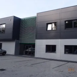 Nowoczesny budynek biurowo-magazynowy z szarymi panelami elewacyjnymi, poziomymi żaluzjami nad wejściem i zaparkowanym srebrnym SUV-em na kostce brukowej.