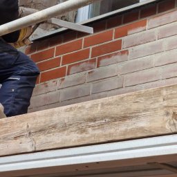 Wood House Group - Pracownik w kasku i osłonie twarzy na rusztowaniu, odnawia ceglany mur przy oknie. Część cegieł pomalowana na biało, widoczne narzędzia i fragment drewnianej deski.