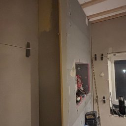 Wood House Group - Montaż płyt gipsowo-kartonowych na ścianie z widoczną instalacją elektryczną w puszce. Widoczne narzędzia i elementy konstrukcyjne. Prace wykończeniowe w trakcie.