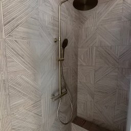 Wood House Group - Wykończona kabina prysznicowa z deszczownicą w odcieniach beżu i brązu. Ściany wyłożone płytkami z geometrycznym wzorem imitującym drewno.