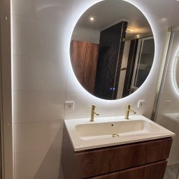 Wood House Group - Nowoczesna łazienka z podświetlanym lustrem LED, białą umywalką nablatową i szafką z drewnopodobnym frontem. Widoczne białe płytki na ścianach i szara podłoga.