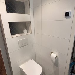 Wood House Group - Nowoczesna toaleta z białymi płytkami, wnękami z ciemną mozaiką w jodełkę, podwieszaną miską WC i szarą podłogą. Minimalistyczny design, czyste linie.