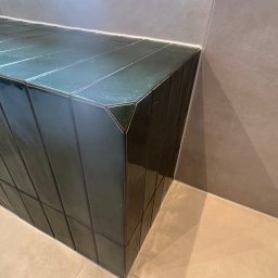 Wood House Group - Zielona, błyszcząca ławka wyłożona płytkami w łazience, z widocznymi fugami i detalami wykończenia, na tle szarej ściany i podłogi. Ujęcie z bliska.