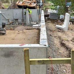Wood House Group - Budowa fundamentów z prefabrykatów betonowych w Wołowie. Widoczny ubijak gruntu, szalunek i wykop. Prace ziemne w toku, przygotowanie do dalszych etapów budowy.