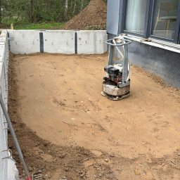 Wood House Group - Przygotowanie podłoża pod budowę: ubita ziemia w betonowych fundamentach, ubijarka wibracyjna i grabie. W tle widoczny dom z szarą elewacją i sterta ziemi.