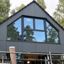 Wood House Group - Nowoczesny dom z ciemną elewacją i dużym trójkątnym oknem, widok z rusztowania. Minimalistyczny design, otoczony zielenią drzew.