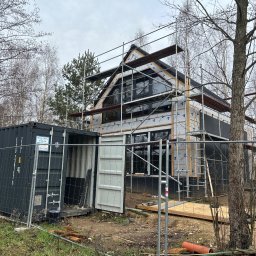 Wood House Group - Wznoszony dom szkieletowy z widocznym ociepleniem, rusztowaniami i kontenerem budowlanym na działce. Widoczne drzewa w tle i pochmurne niebo.