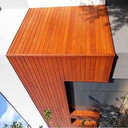 Wood House Group - Nowoczesna elewacja budynku z drewnianą okładziną w ciepłym odcieniu brązu, kontrastująca z białą, teksturowaną ścianą i szarą, chropowatą powierzchnią przy gruncie. Widoczne fragmenty błękitnego...