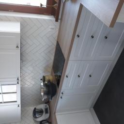 Wood House Group - Kuchnia z białymi szafkami z frezowanymi frontami i okrągłymi uchwytami, blat z jasnego drewna, ściana wyłożona białymi płytkami ułożonymi w jodełkę, widoczny sprzęt AGD na blacie.