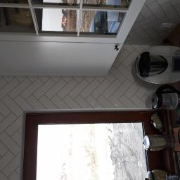 Wood House Group - Ukończona kuchnia z białymi, ułożonymi w jodełkę płytkami na ścianie, białymi szafkami i drewnianym oknem, widok z boku na blat z urządzeniami kuchennymi.