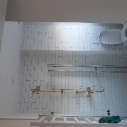 Nowoczesna łazienka z białymi, prostokątnymi płytkami ułożonymi pionowo, złotą armaturą prysznicową, szklanym panelem prysznicowym i wiszącą toaletą. Fragment dekoracyjnej ściany z czarno-białą...