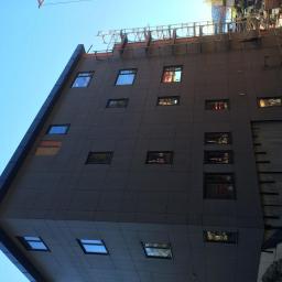 Wood House Group - Wielopiętrowy budynek z ciemnoszarą elewacją z paneli, widoczny z dołu pod kątem, z rusztowaniami i dźwigiem w tle.