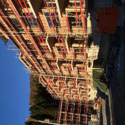 Wood House Group - Budowa wielopiętrowego budynku w trakcie wznoszenia, widoczne rusztowania, elementy konstrukcyjne i żuraw na tle lasu i błękitnego nieba.