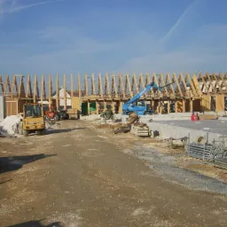 Wznoszenie konstrukcji drewnianej budynku na fundamencie betonowym, widoczny dźwig, betoniarka i sprzęt budowlany na placu budowy w słoneczny dzień.