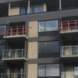 Wood House Group - Wielopiętrowy budynek z balkonami z szklanymi balustradami, na niektórych balkonach widoczne czerwone elementy zabezpieczające, z boku budynku przymocowana drabina rusztowaniowa.